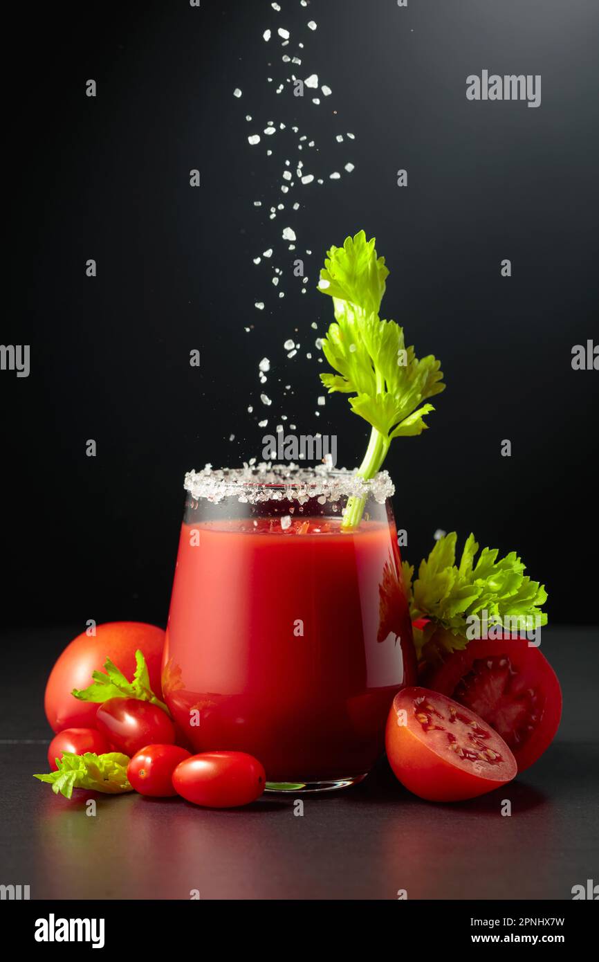 Sea salt is poured into a glass of tomato juice with celery. The glass of juice and fresh vegetables are on a black background. Stock Photo