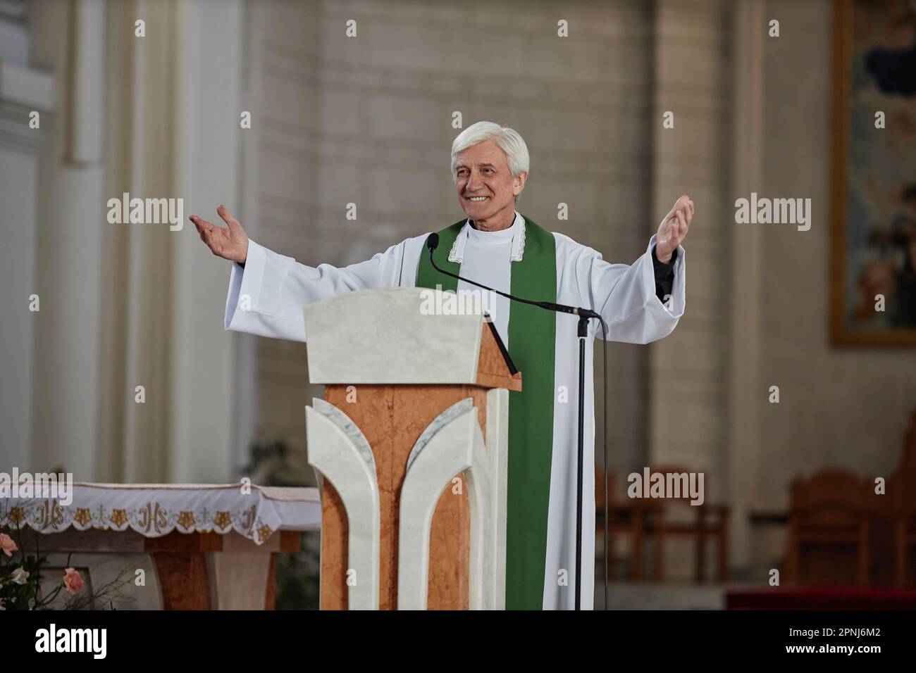 Smiling senior priest in formalwear greeting people in church while