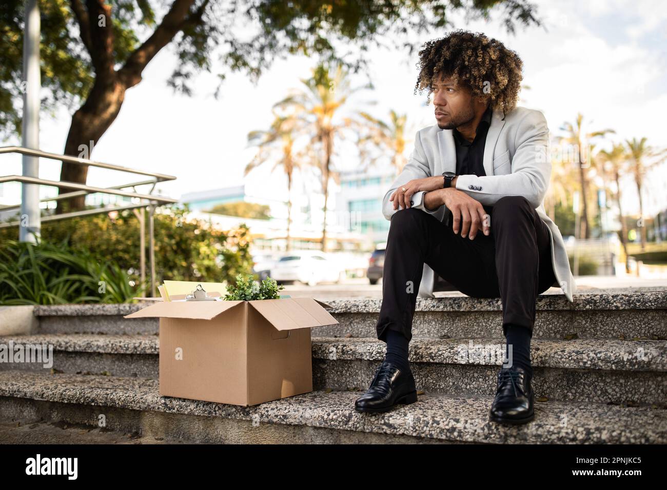 Crisis and unemployment. Upset black middle aged office worker sitting with box of personal stuff on stairs outdoors Stock Photo