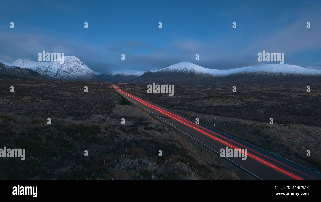 Panorama of a road leading through the Scottish Highlands of Glen Coe with car light trail, snowcapped mountains and valley. Scotland Stock Photo