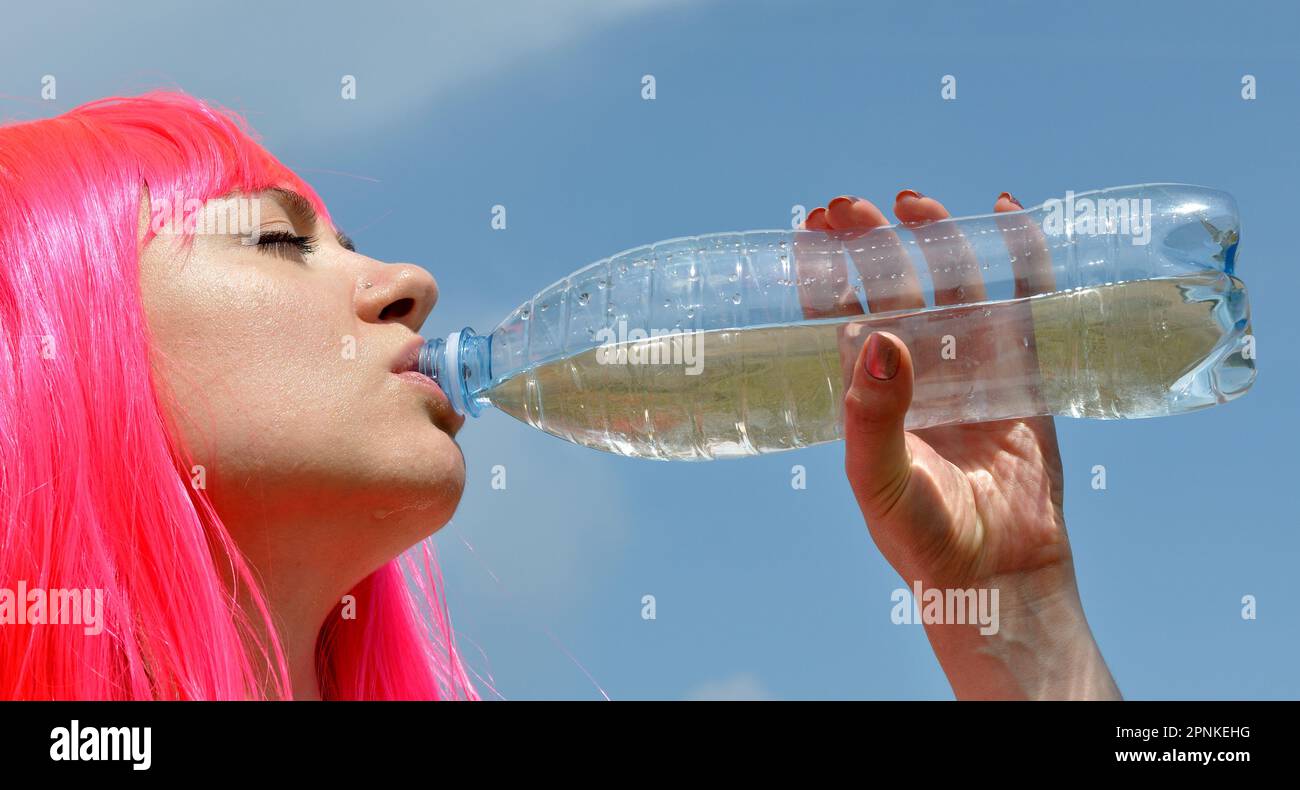 young attractive woman with pink hair drinks cold water in hot day on blue sky background Stock Photo