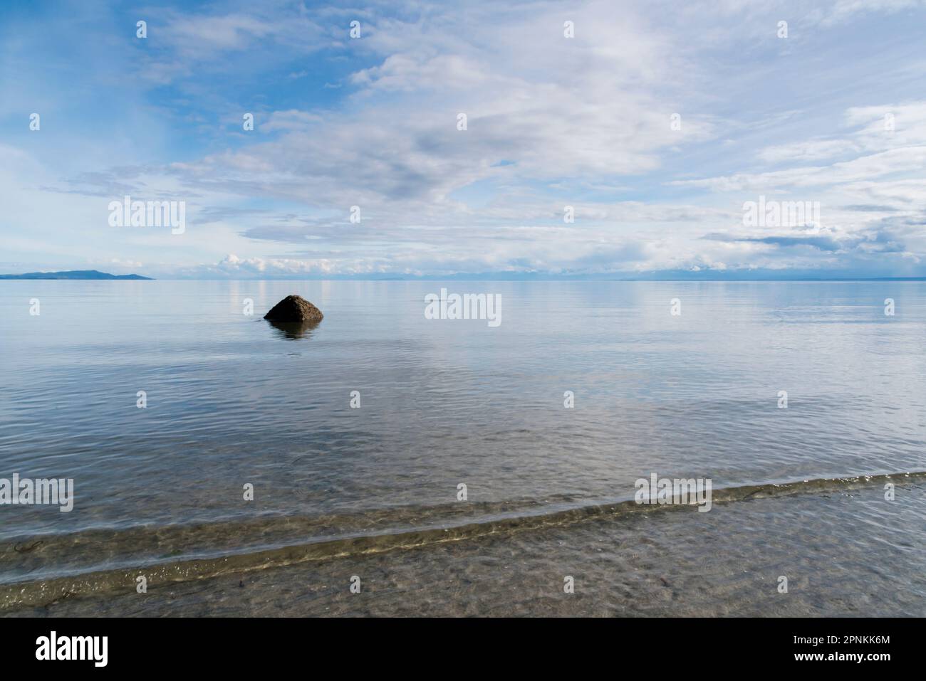 A boulder emerges from calm water on a sunny afternoon at low-tide as gentle waves break over the white sand beach at Savary Island, British Columbia Stock Photo