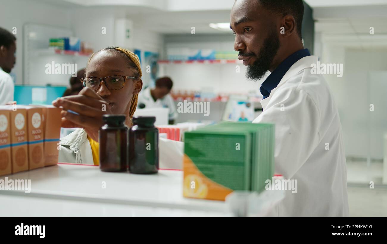 African american man recommending vitamins to woman in drugstore, talking about healthcare and supplements. Retail store worker shoing box of pills to female client, choosing drugs. Stock Photo