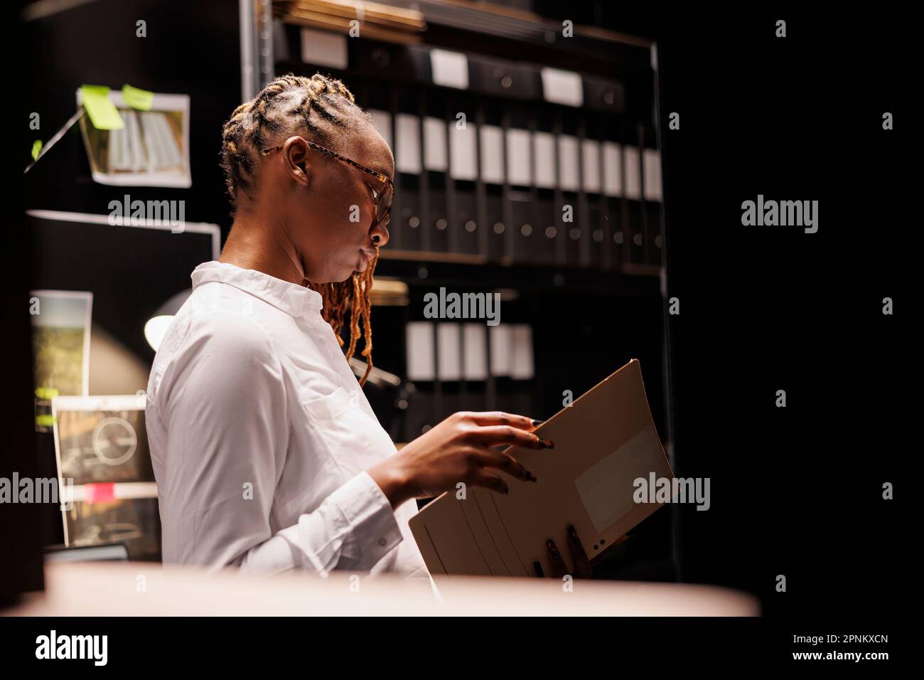Thoughtful african american woman detective reading criminal investigation documentation in agency office room. Law enforcement professional studying criminologist report at night Stock Photo