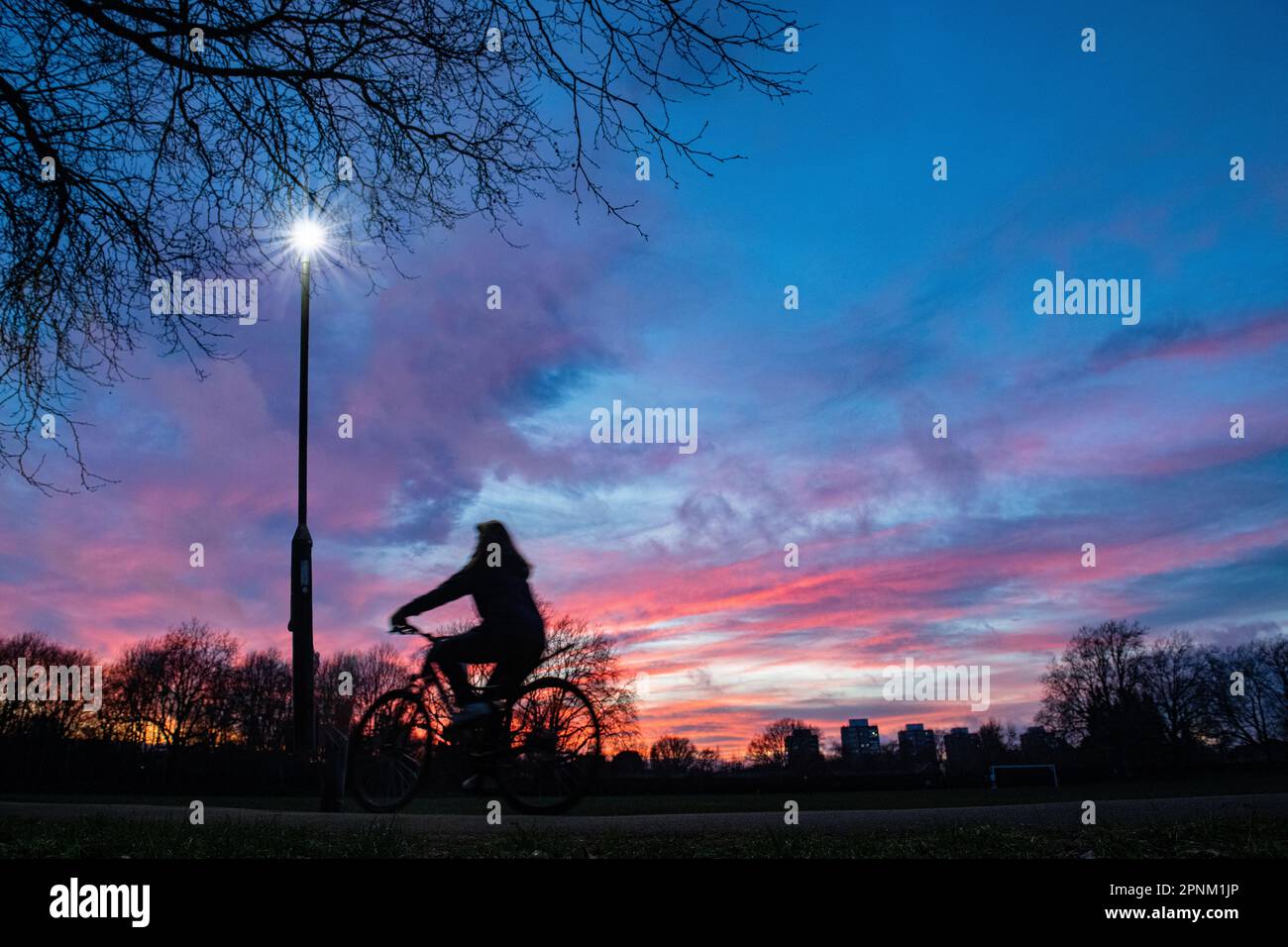 A cyclist rides through a London park at sunset under  street light Stock Photo