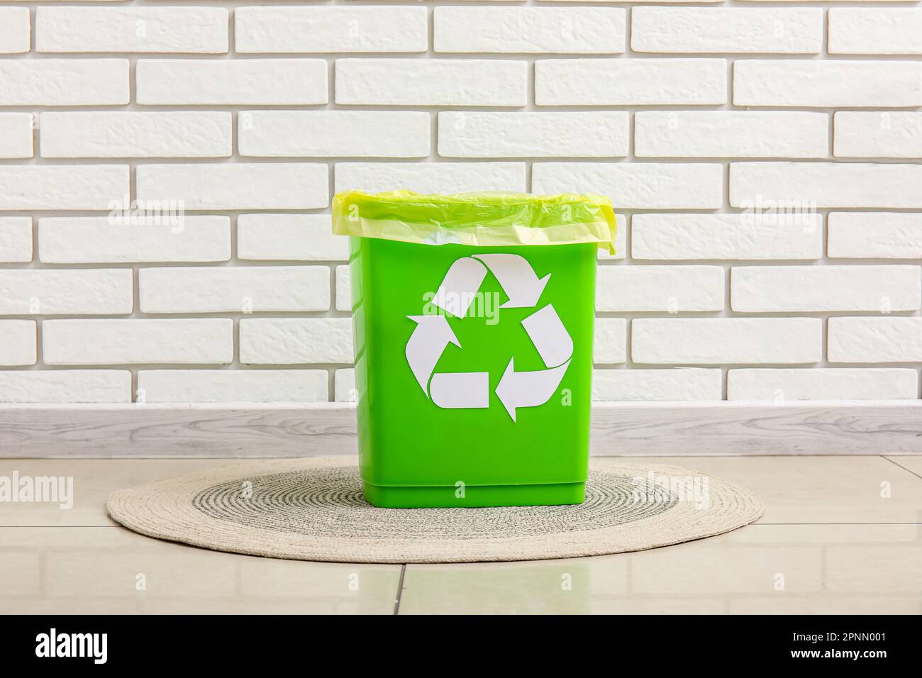 Trash bin with recycling symbol on rug near white brick wall Stock