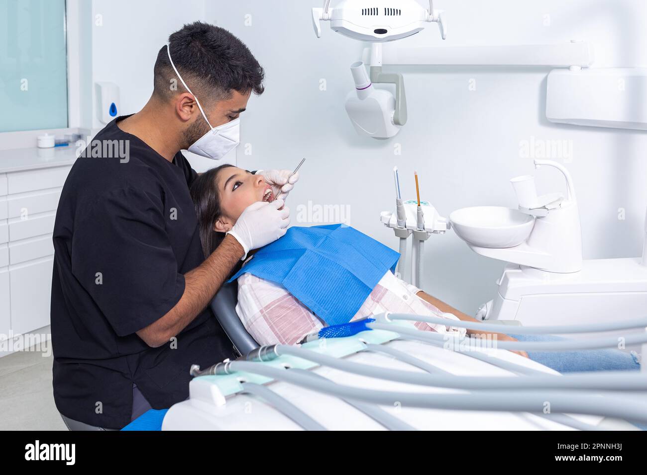 Male dentist in gloves and face mask using mirror to inspect teeth of teen patient during check up in light office of dental clinic Stock Photo