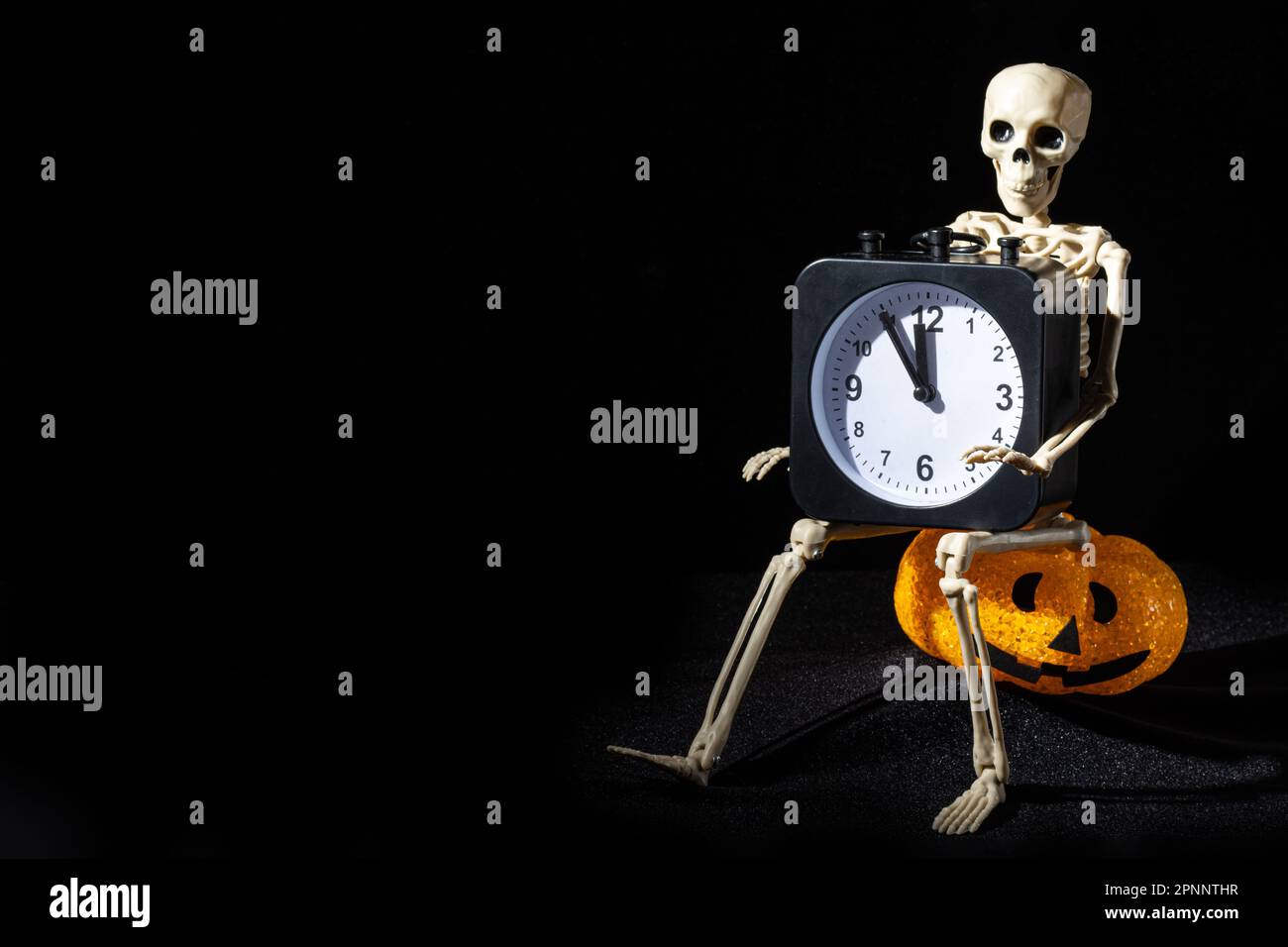 A skeleton sitting on a letter-shaped lamp with a grin and holding an alarm clock on a black background. Time of the Dead. Stock Photo