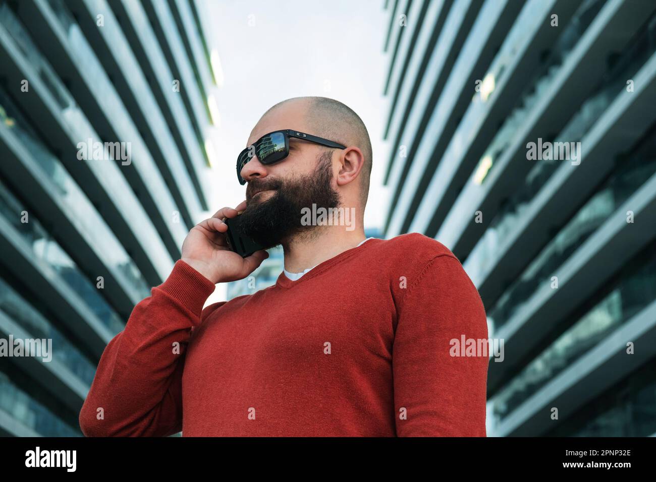 Bearded man in sunglasses talking on mobile phone outdoor the modern office buildings. IT guy or freelancer using mobile phone. Close up Stock Photo