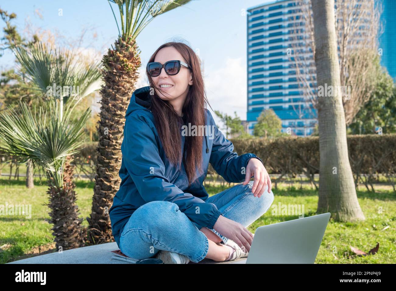 Young attractive cheerful woman in sunglasses working remotely in the park using laptop and smiling. Remote work, freelance, lifestyle concept Stock Photo