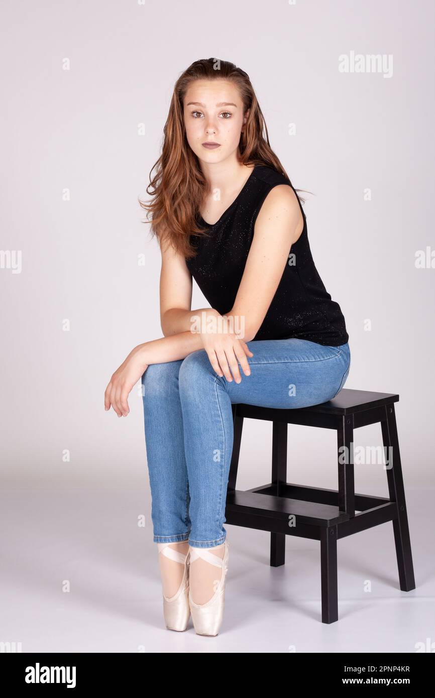 En Pointe resting on stool legs together unemotional en pointe Beautiful teen girl sitting straight looking at the camera, hand to chin with an intima Stock Photo