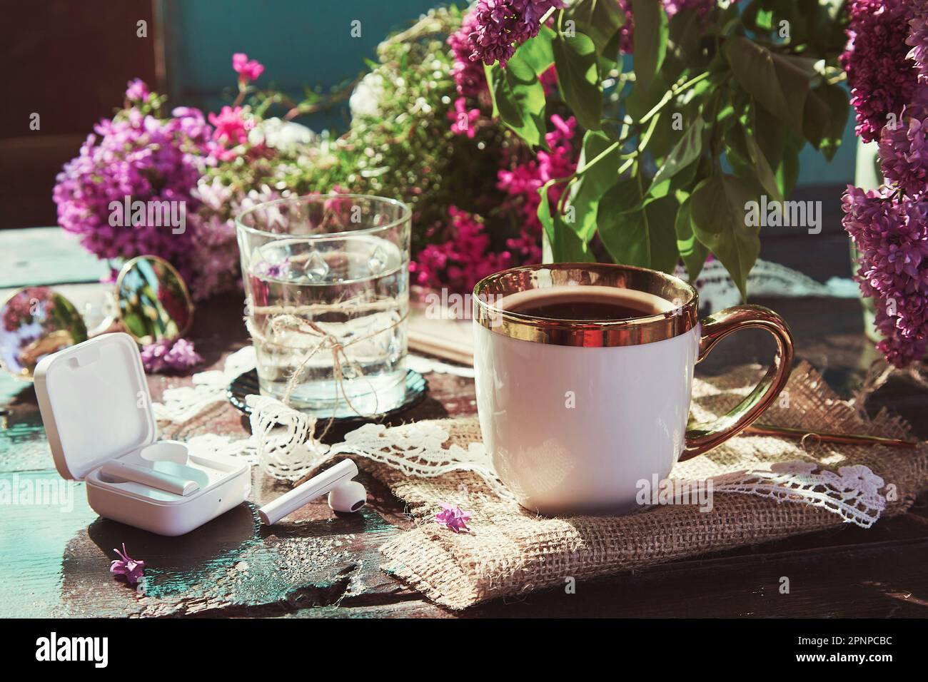 Coffee cup and glass of water on the table. Wireless headphones, listen to music, audiobook concept. Springtime lilac flowers outside Stock Photo