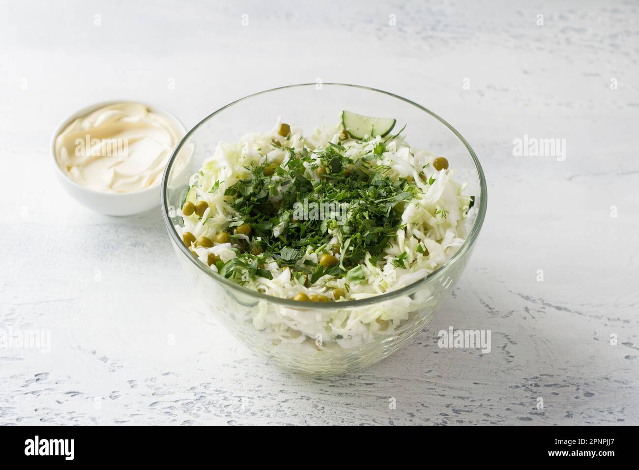 Glass bowl of cabbage salad with cucumbers, green peas, chopped herbs and mayonnaise dressing. Cooking delicious homemade salad. Step-by-step instruct Stock Photo