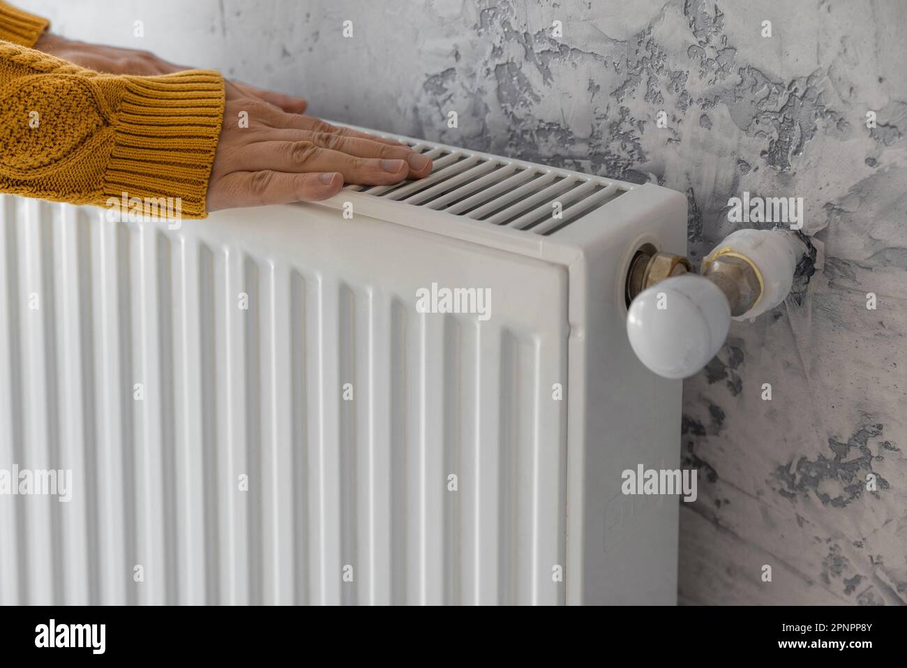 Man in yellow sweater warming his hands on the heater at home during cold winter days. Male getting warm up his arms over radiator. Concept of heating Stock Photo
