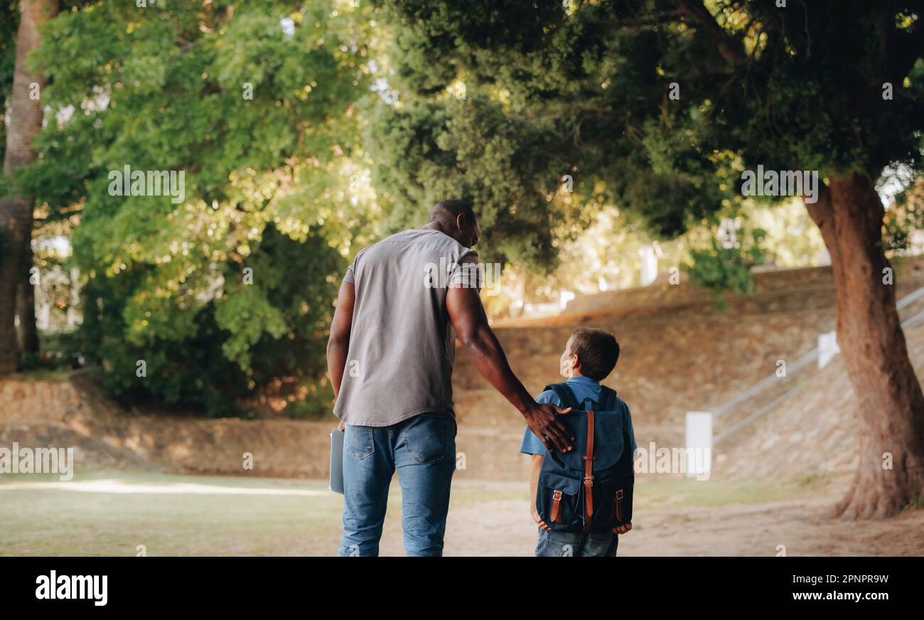 Child mentorship in primary school. Rearview of a male teacher talking and walking with a young school kid outside. Providing support and encouragemen Stock Photo