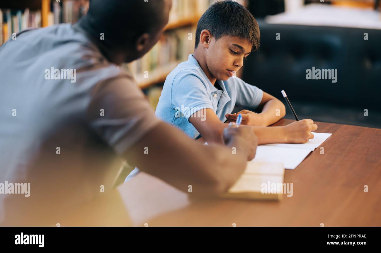 Child counselling in elementary school. Young school boy writing in a journal while sitting in a library with an educator. Primary school student havi Stock Photo