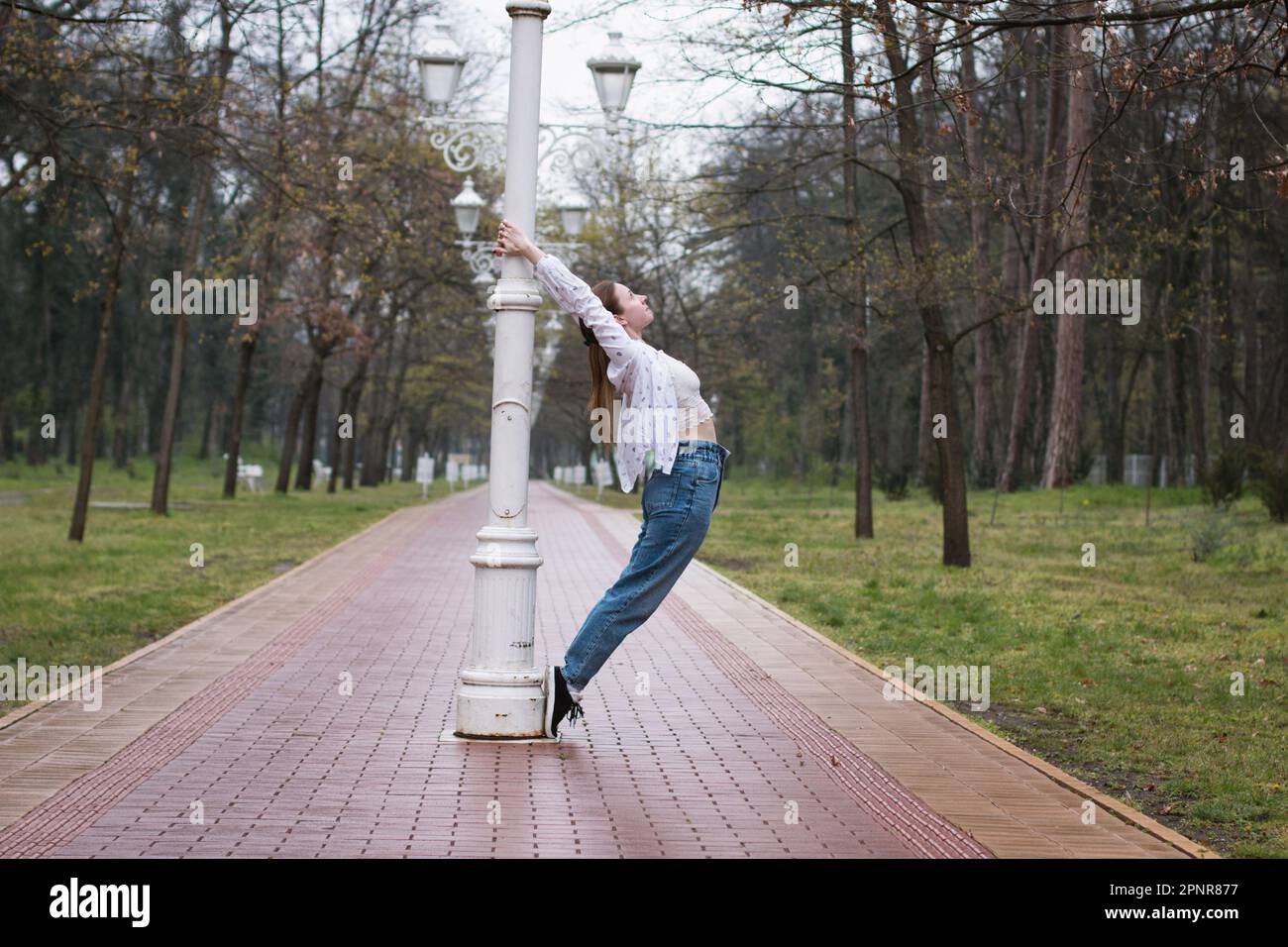 Young woman dancing around the light pole outdoors in the park, in rainy weather. Stock Photo