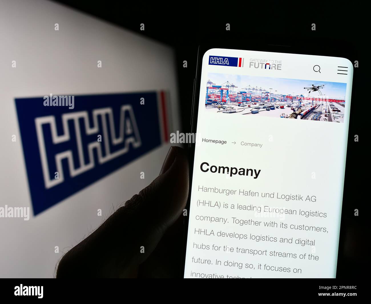 Person holding smartphone with webpage of company Hamburger Hafen und Logistik AG (HHLA) on screen with logo. Focus on center of phone display. Stock Photo