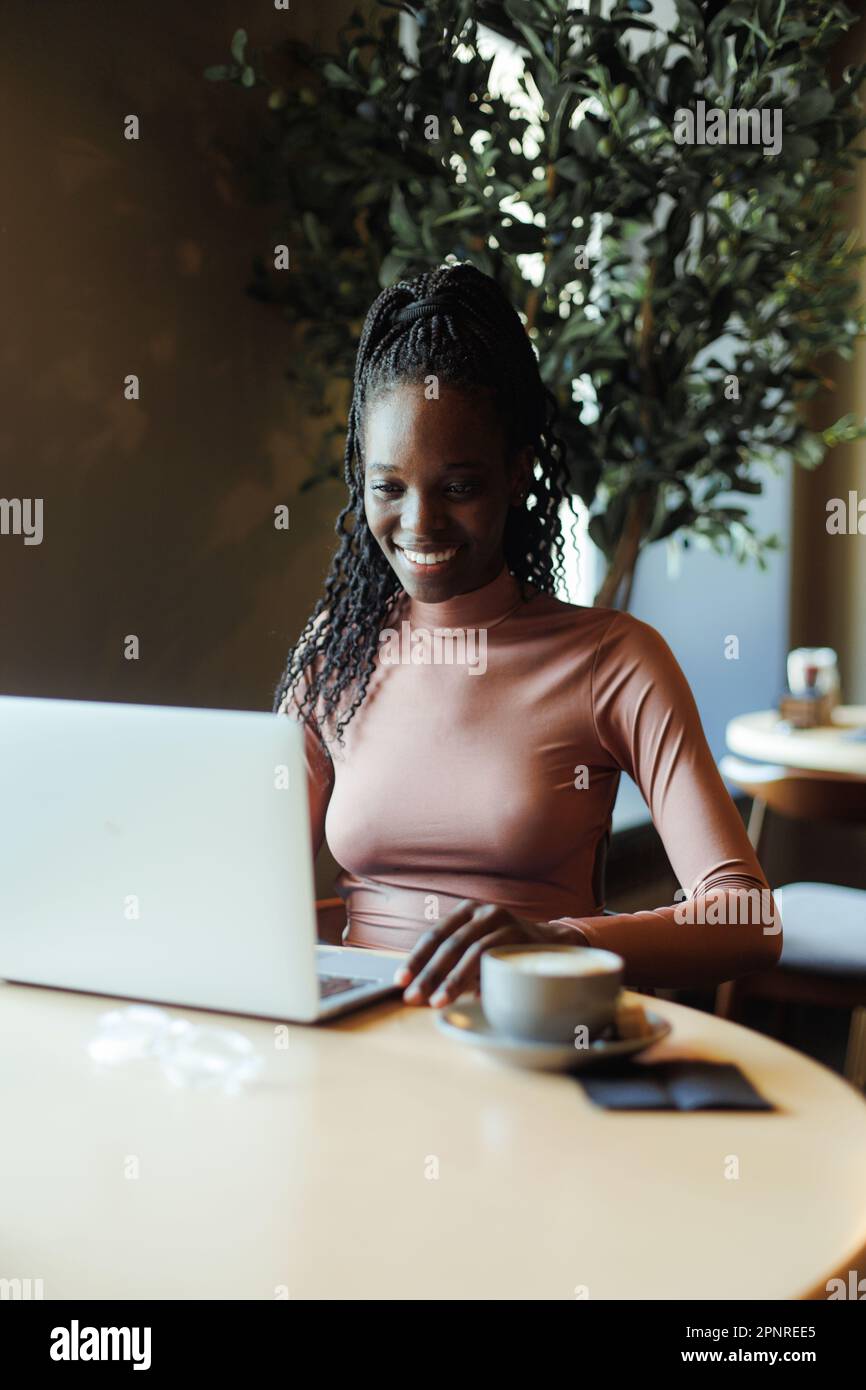 Portrait of young joyful African-American woman sitting at beige table in cafe near laptop, looking at screen, chatting. Stock Photo