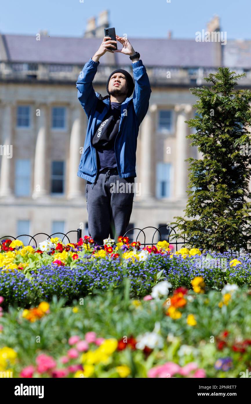 Bath, UK. 20th April, 2023. With Bath's famous Royal Crescent in the background a man is pictured taking a photograph of the colouful flower beds in Royal Victoria Park. Forecasters are predicting that cooler and more overcast weather is to come in the next few days. Credit: Lynchpics/Alamy Live News Stock Photo
