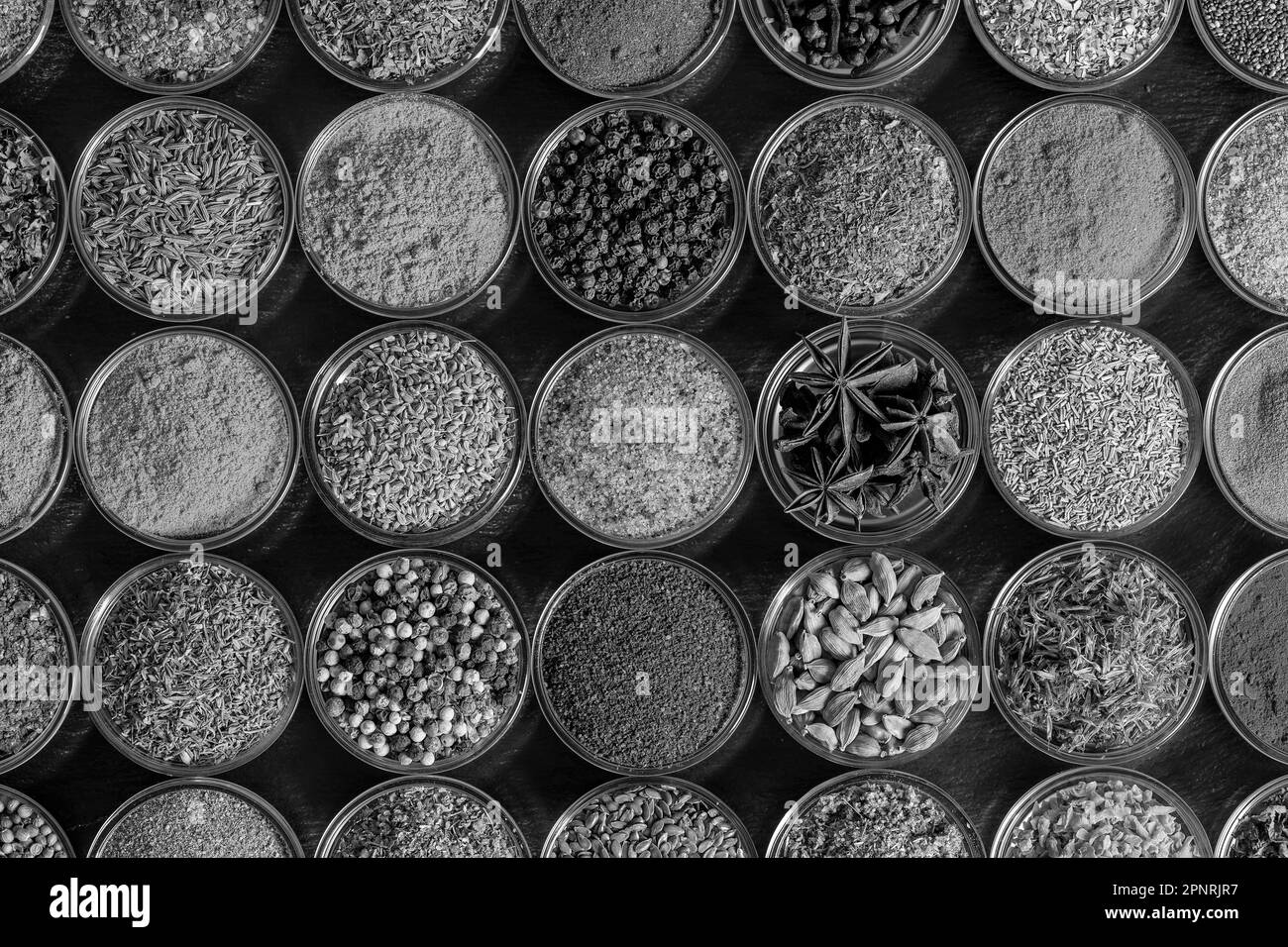 Assortment of aromatic spices, seeds and dry herbs for cooking food on background in small glass bowls, close up, top view, black and white Stock Photo