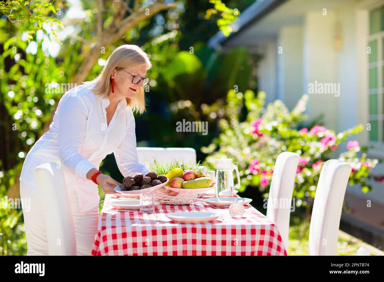 Woman setting table outdoors. Garden summer fun. BBQ in sunny backyard. Senior lady cooking lunch. Party decoration. Female with fruit outdoor. Stock Photo