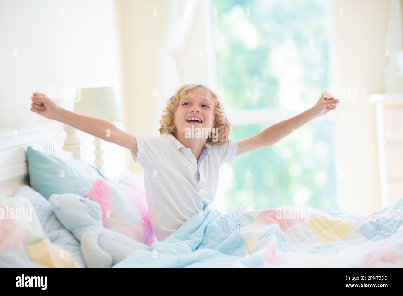 Child in bed on sunny morning. Kid waking up in white bed at big window
