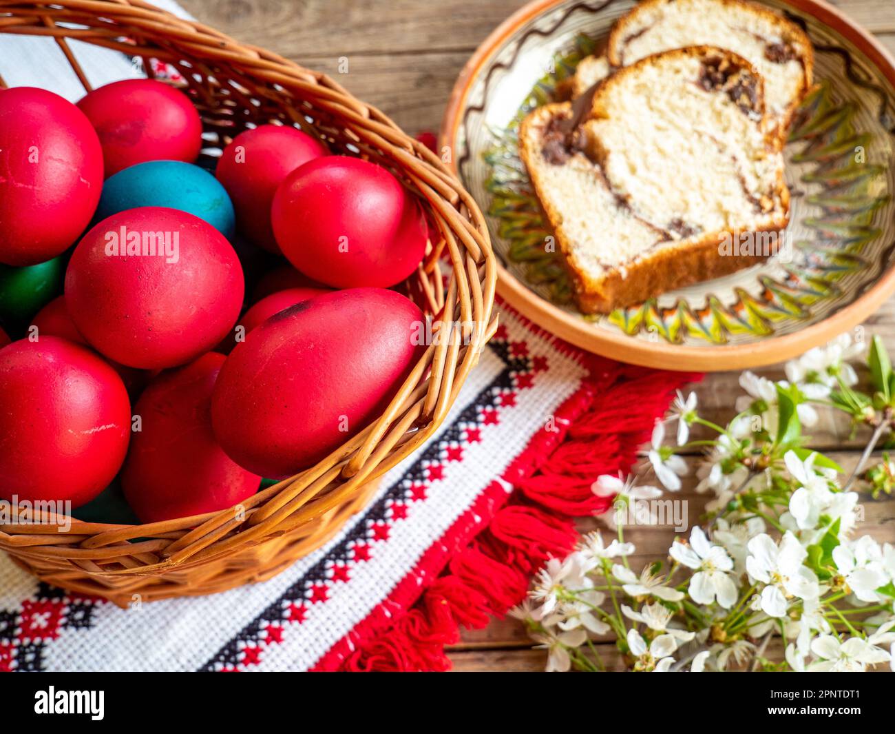 red Easter eggs next to flowering branches and plate with sweet sponge cake or cozonac on wooden table Stock Photo