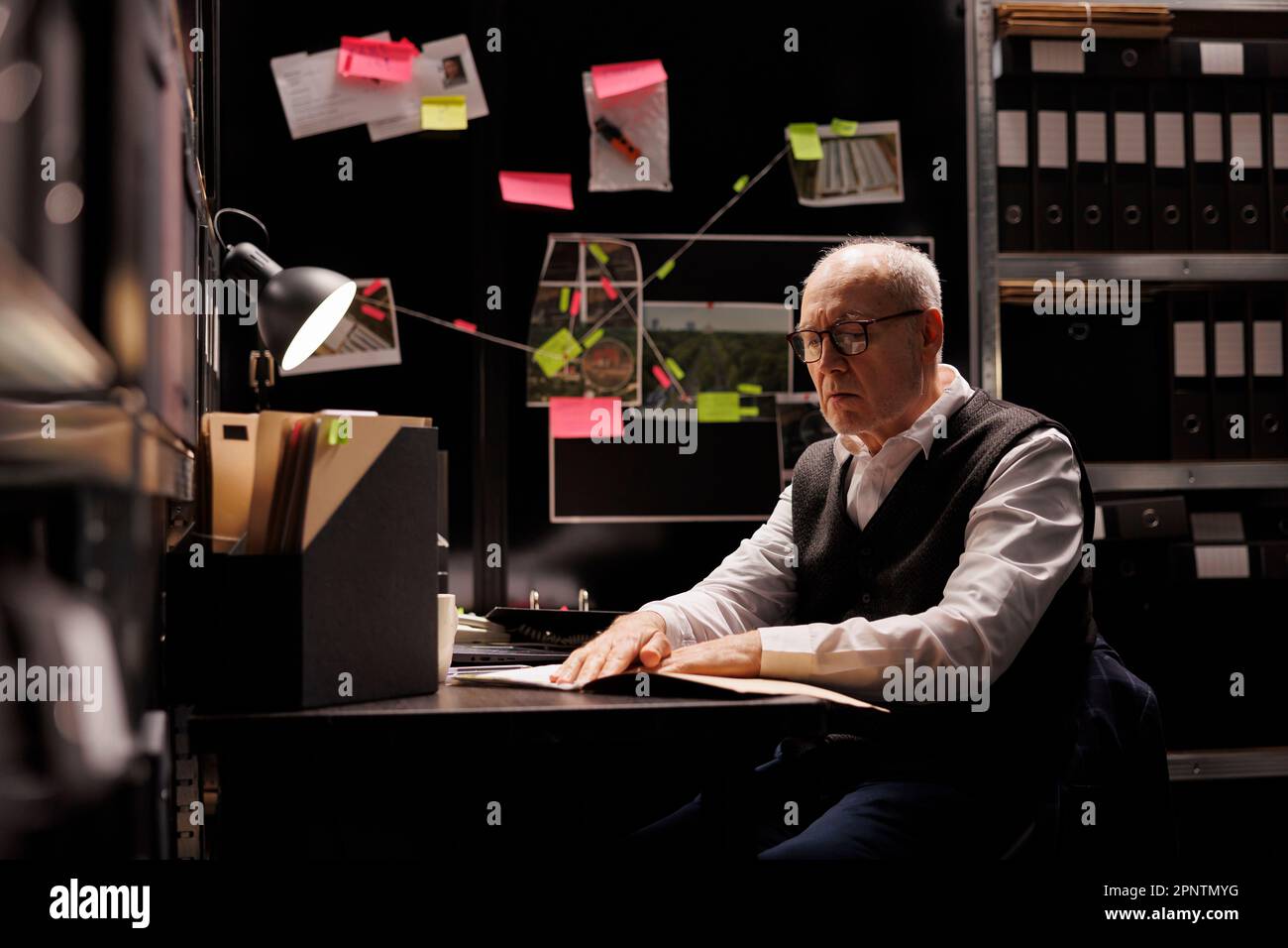Senior private investigator analyzing criminology document, working overtime at criminal case in arhive room. Elderly police officer checking victim files, looking at mysterious suspect report Stock Photo