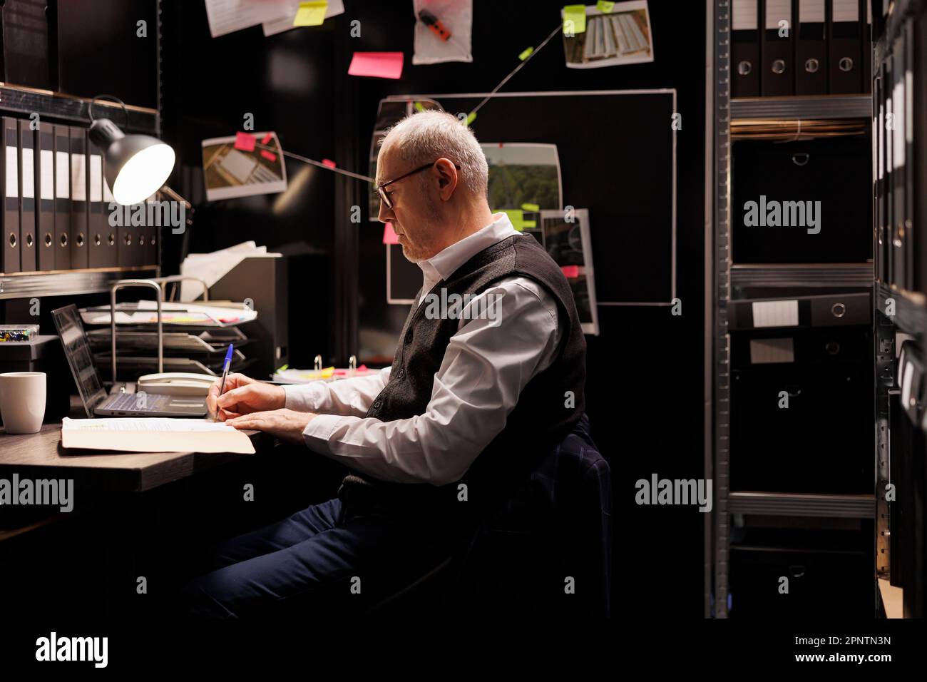 Elderly private detective writing missing person case report, working late at night at criminal case in arhive room. Old police officer analyzing criminology files, planning startegy to catch suspect Stock Photo