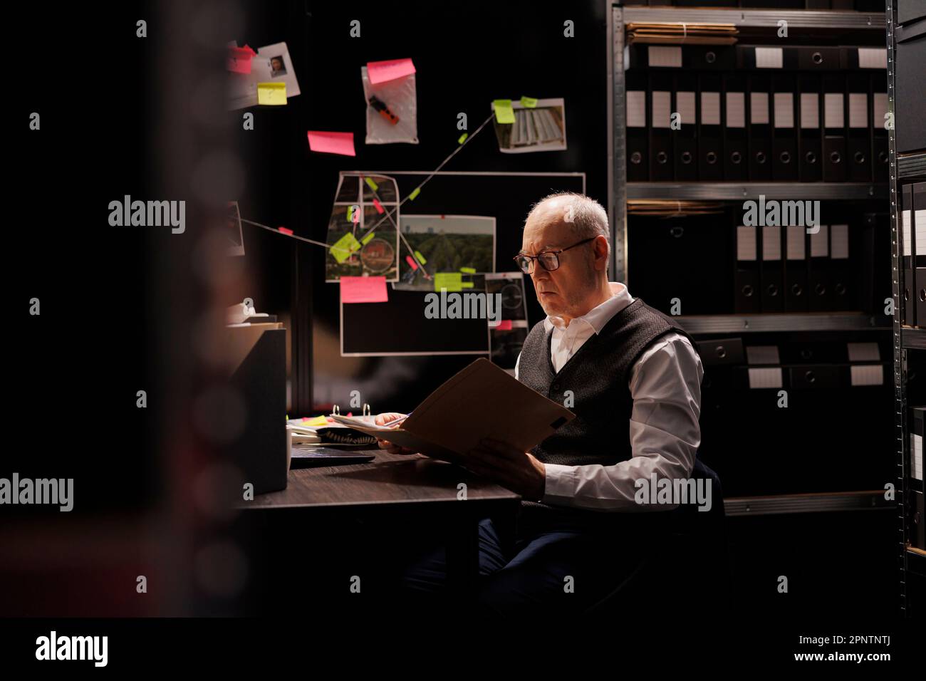 Senior police officer looking at criminal investigations files, working overhours at mysterious suspect profile in arhive room. Elderly private detective analyzing crime scene evidence Stock Photo