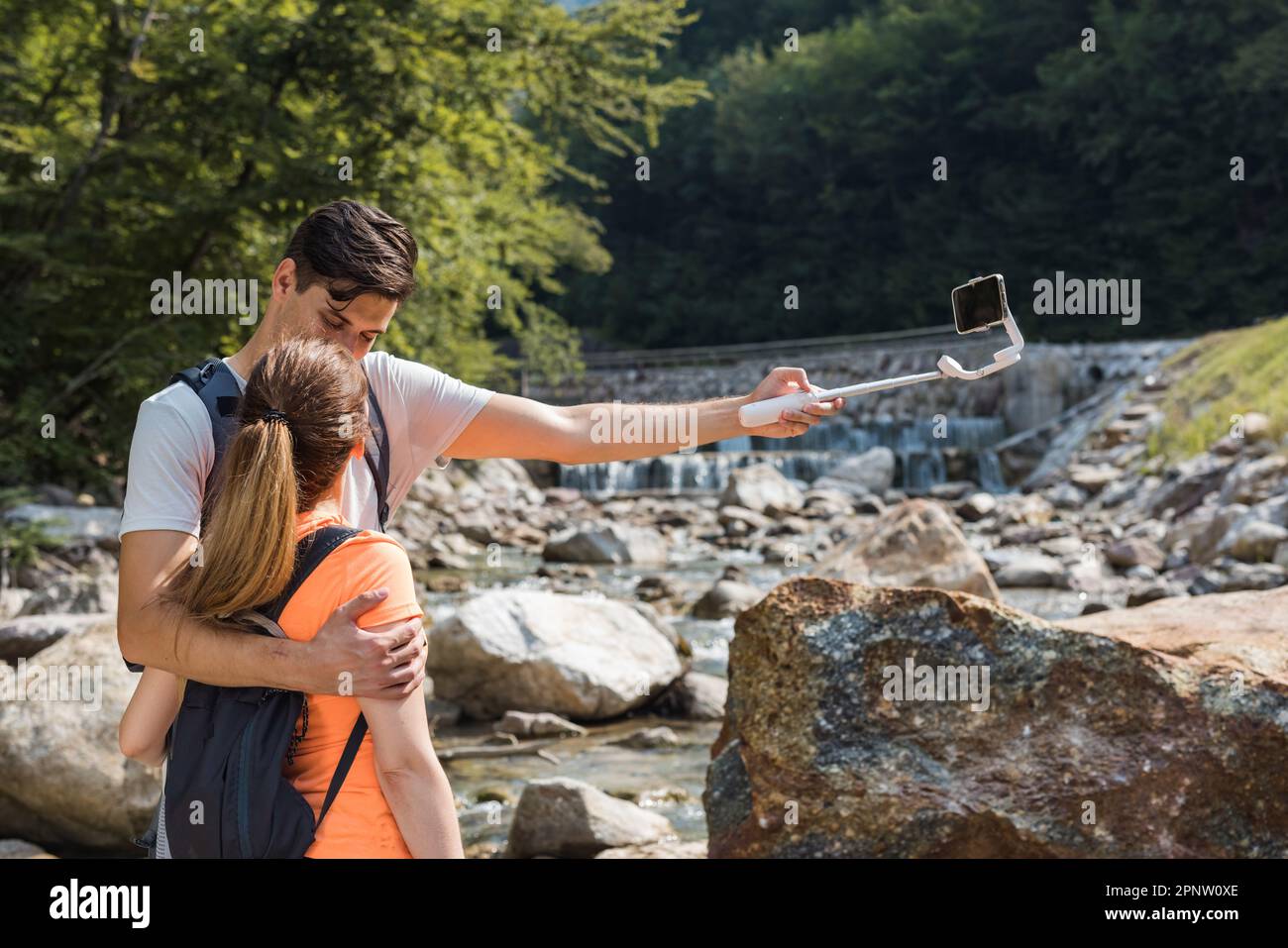 Handsome young couple standing in nature near the mountain river, making selfie vlog videos using a smartphone and selfie stick Stock Photo