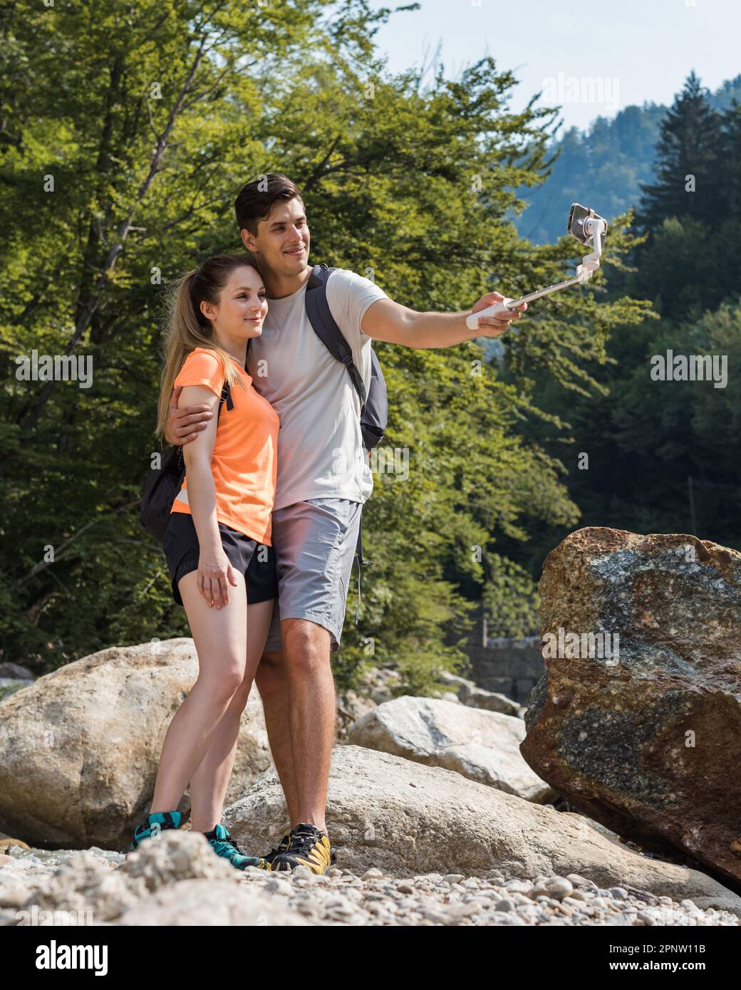 Handsome young couple standing in nature near the mountain river, making selfie vlog videos using a smartphone and selfie stick Stock Photo