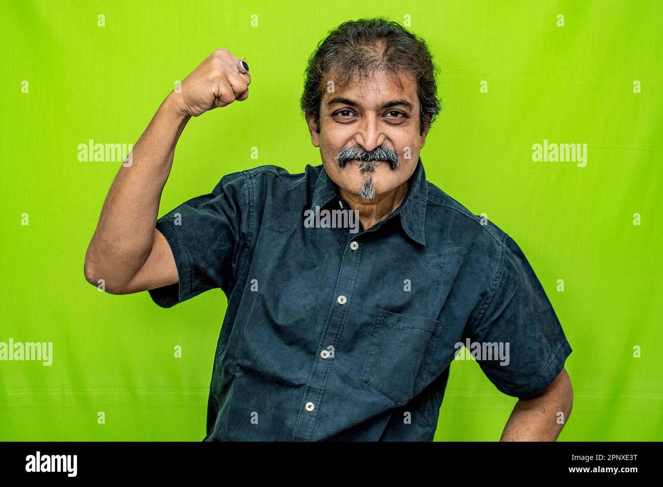 A well-dressed corporate man in a black shirt stands against a green screen, appearing to celebrate a win as he raises his right hand in a closed fist Stock Photo