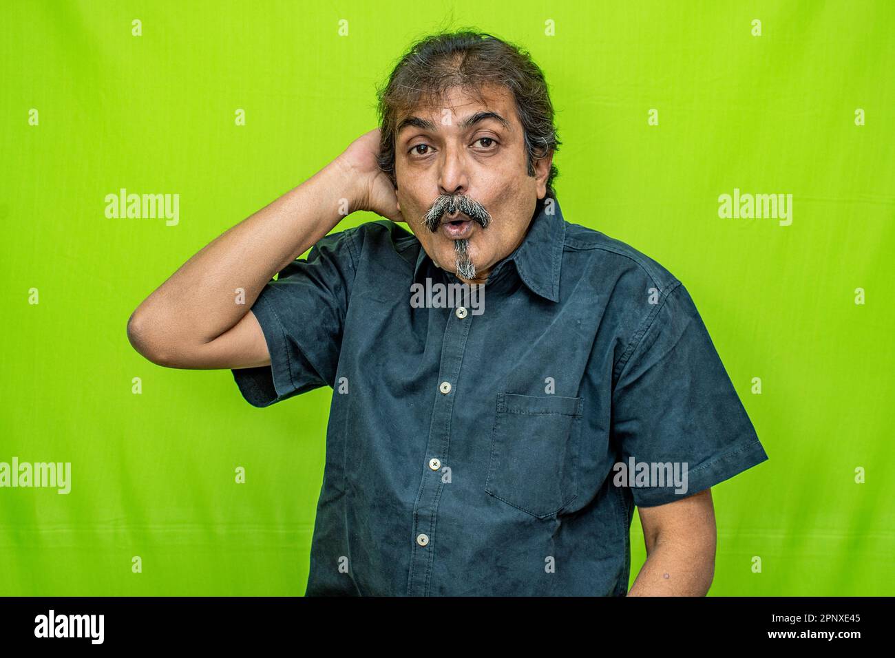 A well-dressed businessman in black shirt stands against a green screen. He touches his head with a confused expression, realizing he forgot something Stock Photo