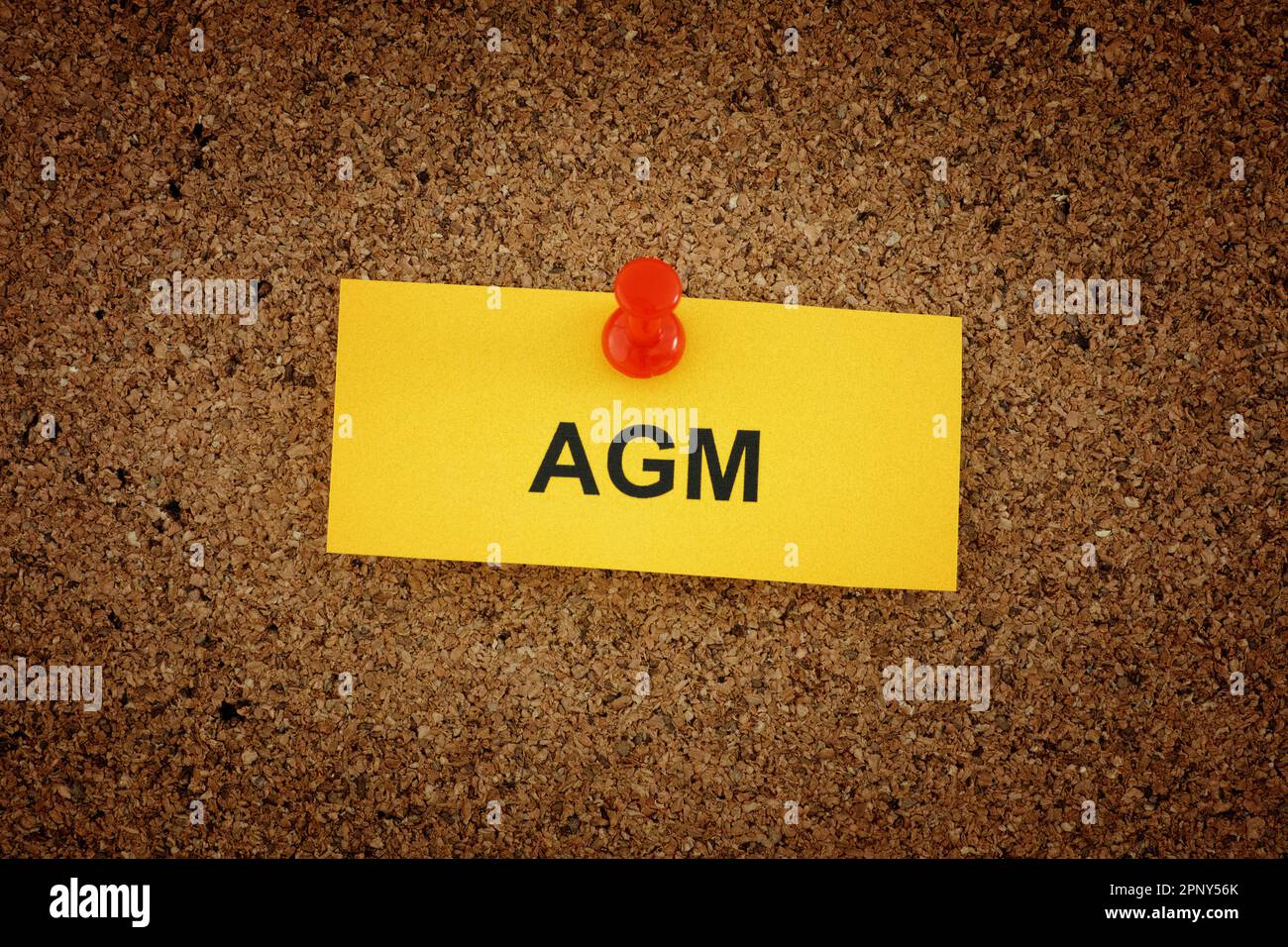 A yellow paper note with the words AGM (Annual General Meeting) on it pinned to cork board. Close up. Stock Photo