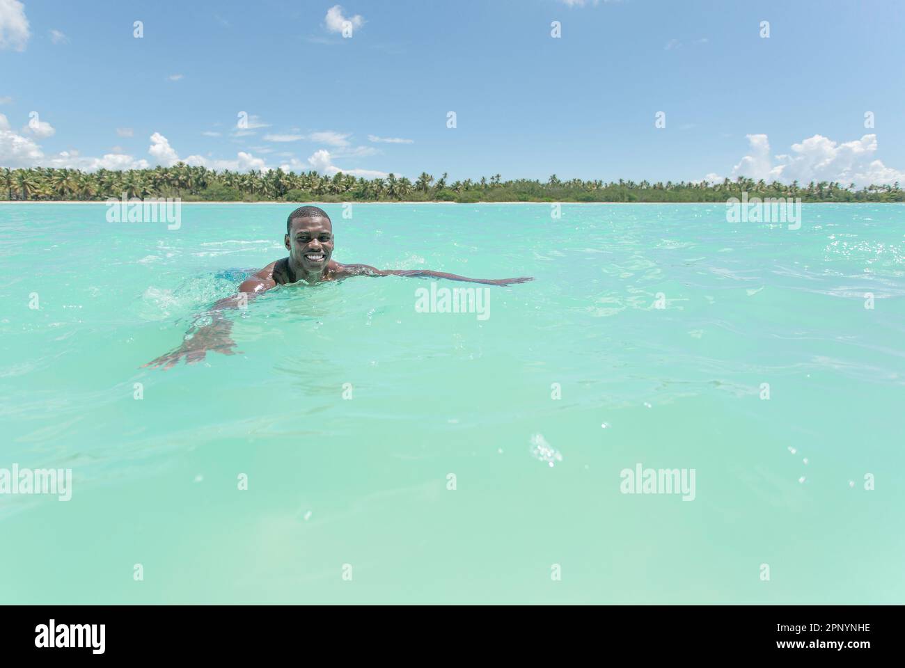 A young black man can be seen swimming and enjoying the crystal-clear turquoise waters of the Caribbean sea. In the background, a lush green island an Stock Photo