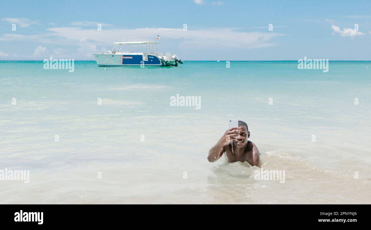 A young black man is seen lying on the shore of the ocean, smiling and taking a selfie with a boat in the background. The sky is a beautiful blue with Stock Photo