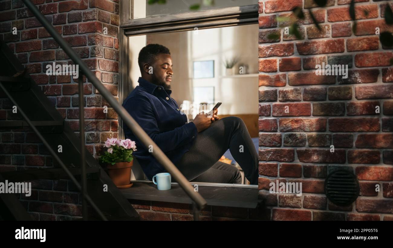 Black Authentic Man Using Smartphone in his Flat While Sitting on Bedroom Windowsill. African American Male Posting Photos on Social Media and Using Stock Photo