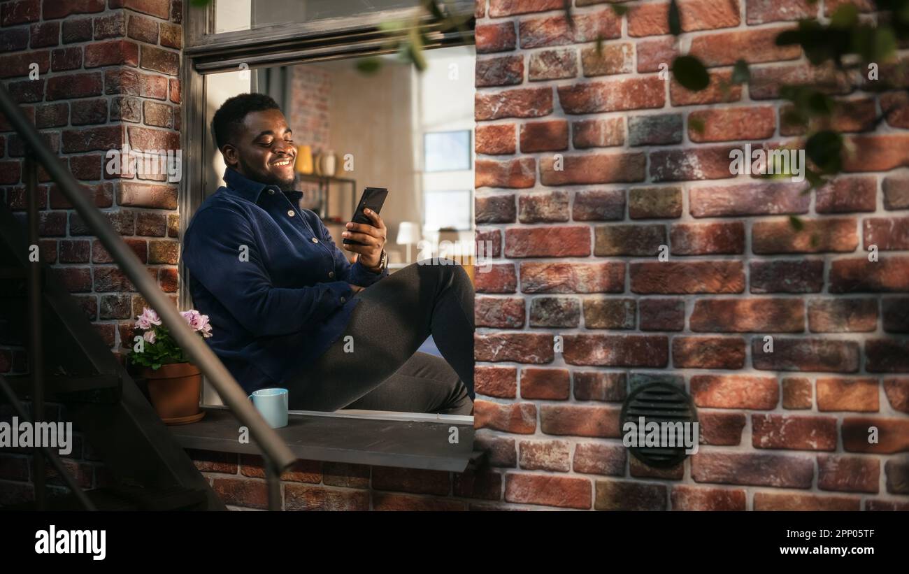 Black Authentic Man Using Smartphone in his Flat While Sitting on Bedroom Windowsill. African American Male Posting Photos on Social Media and Using Stock Photo