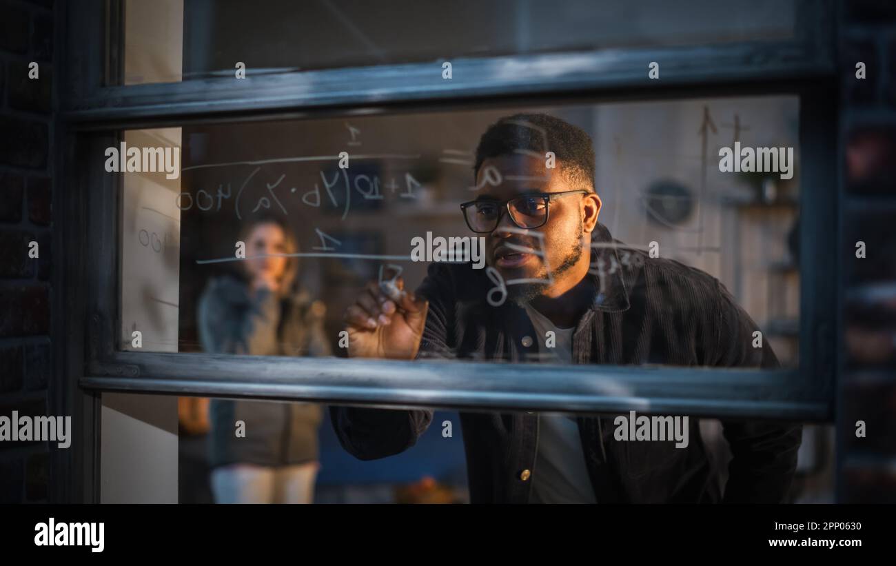Black Young Man Explaining a Mathematical Equation on a Window at Home to a Female Friend by Using Erasable Glass Markers. Two Multiethnic Students Stock Photo