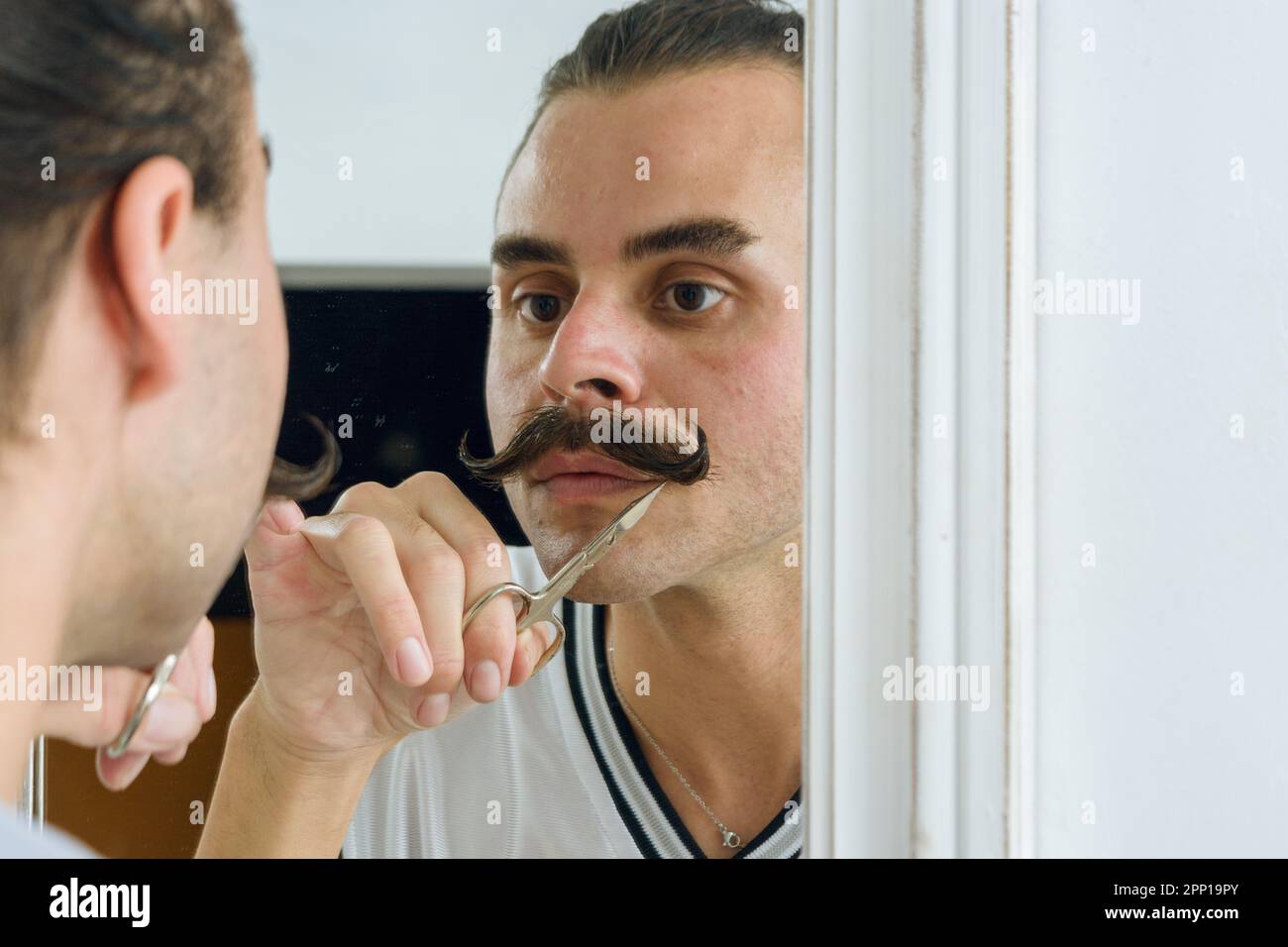 reflection in the mirror of a non-binary Argentinian Hispanic Latino person, concentrated fixing his mustache with scissors Stock Photo