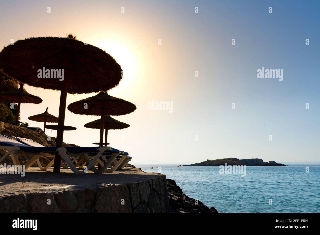 Umbrellas and loungers at sunrise next to the rocky coast Stock Photo