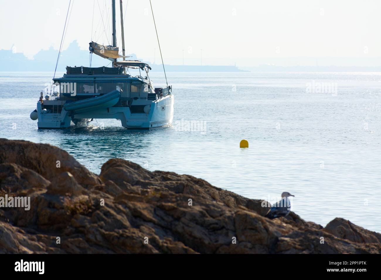 Catamaran anchored off the rocky coast of Ses Illetes with the port of Palma in the background in the mist Stock Photo