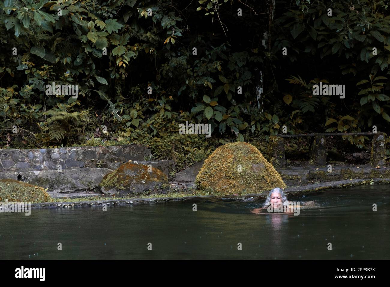 Gentle rain falls as a senior woman swims in a jungle rock pool fed by Chorro Macho near El Valle Anton in Panama Stock Photo