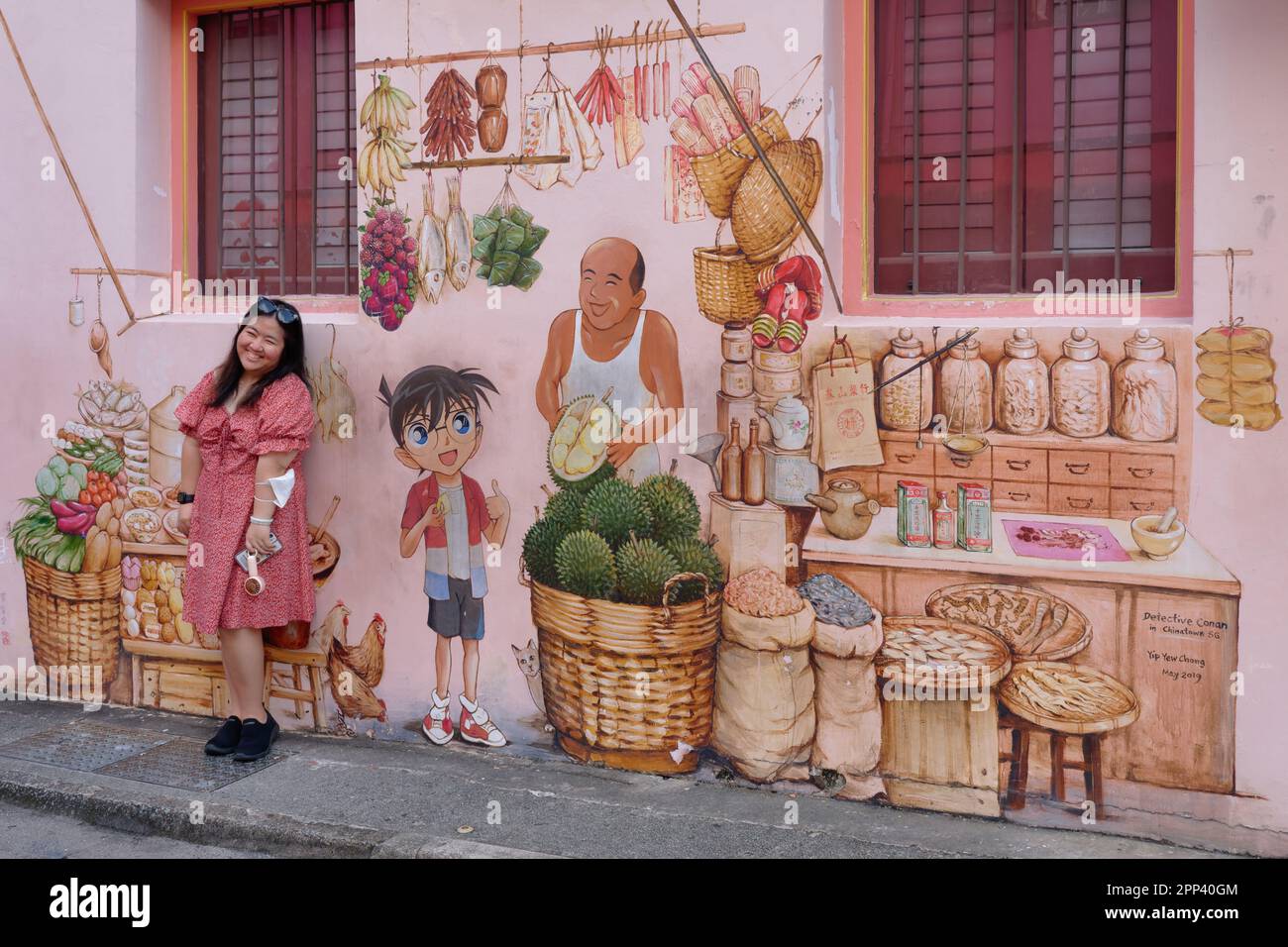 A female Thai tourist poses in front of a mural by artist Yip Yew Chong in Chinatown, Singapore, depicting a Chinese vendor of durians and other foods Stock Photo
