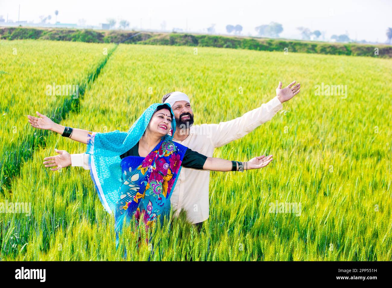 Happy young indian farmer couple standing with open arms at wheat ...