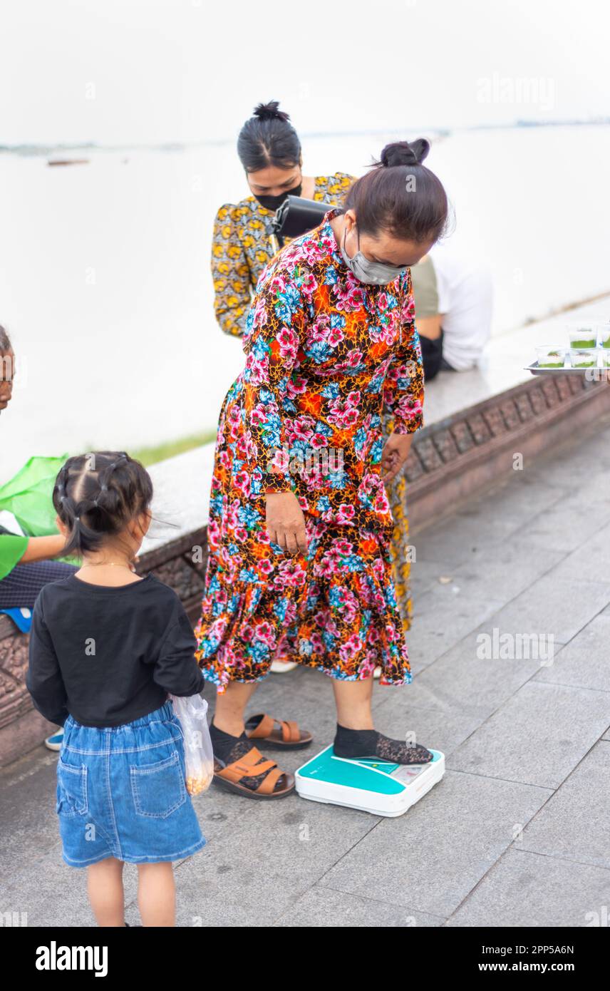 Phnom Penh,Cambodia-January 4th 2023: Khmer woman,with her family,checks her weight on some bathroom scales,along Sisowath Quay boulevard,at sunset. Stock Photo