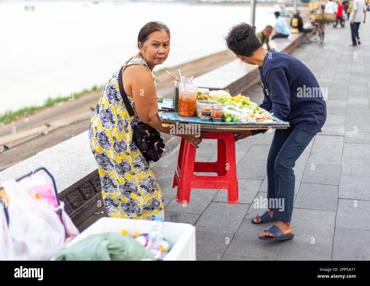 Phnom Penh,Cambodia-January 4th 2023: A woman vendor gets assistance,lifting her tiny fruit display to a different location,to sell fruit snacks along Stock Photo
