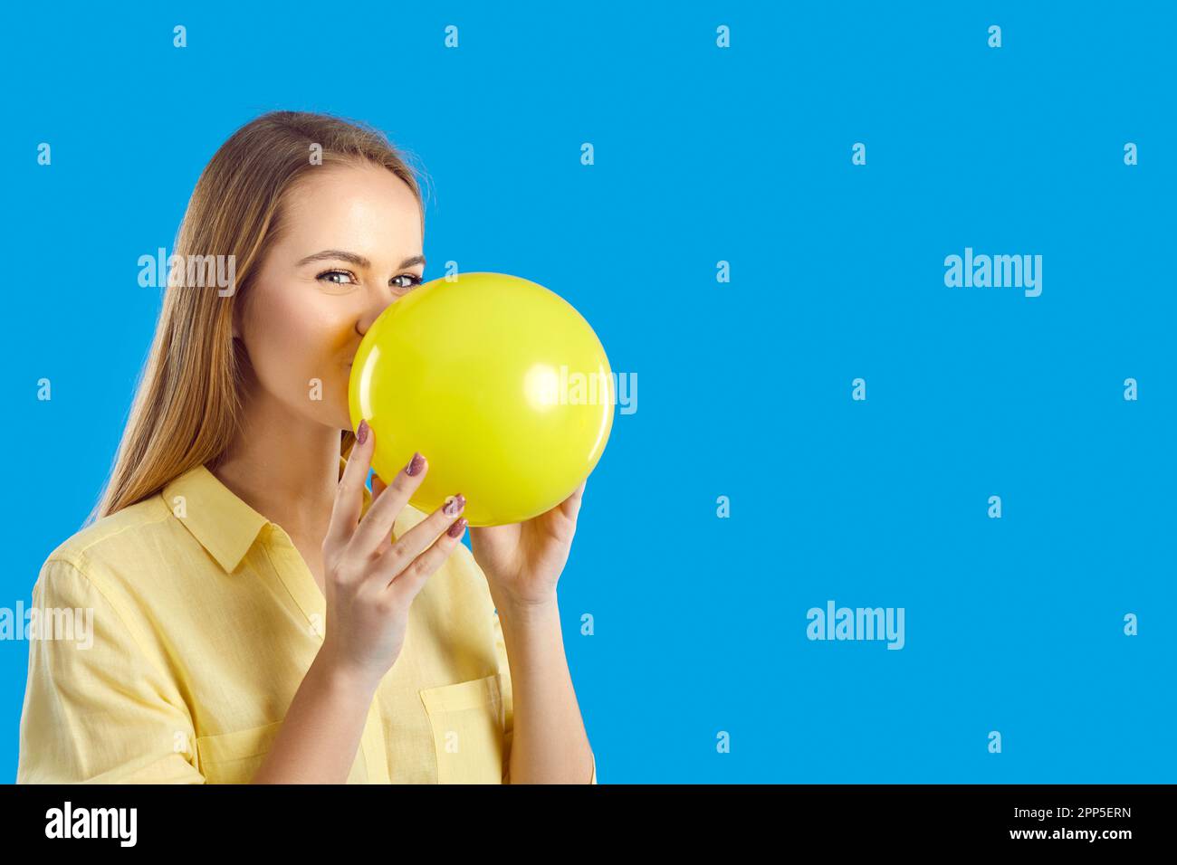 Happy beautiful young woman blowing up yellow balloon on blue copy space background Stock Photo